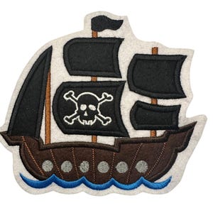 Könnte beinhalten: Ein Piratenschiff-Applikation in Braun, Schwarz und Weiß, mit einem blauen Wellendetail am unteren Rand. Das Schiff hat eine Totenkopf-Flagge und ist aus Filz oder einem ähnlichen Stoff gefertigt.