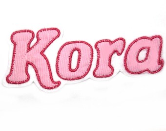 May include: Pink felt name patch with the name "Kora" in a white outline.