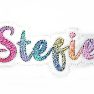 Puede incluir: Un parche bordado colorido con el nombre "Stefanie" en un esquema de colores arcoíris. Las letras están cosidas en una fuente cursiva y tienen una textura ligeramente en relieve.