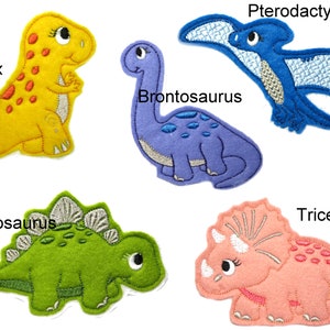 May include: Five felt dinosaur patches, including a yellow T-Rex, a purple Brontosaurus, a blue Pterodactyl, a green Stegosaurus, and a pink Triceratops.