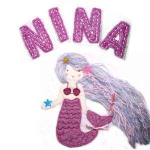 May include: Pink felt appliqu&eacute; of a mermaid with long purple hair, a pink seashell tail, and a blue star in her hand. The name "NINA" is spelled out in pink felt letters above the mermaid.