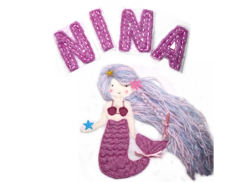 May include: Pink felt appliqu&eacute; of a mermaid with long purple hair, a pink seashell tail, and a blue star in her hand. The name "NINA" is spelled out in pink felt letters above the mermaid.