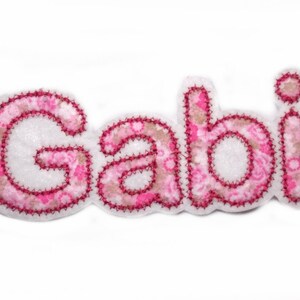 Könnte beinhalten: Rosa Stoffapplikation mit dem Namen "Gabi" in weißen Buchstaben mit rosa floralen Akzenten.