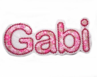 May include: Pink fabric applique with the name "Gabi" in white letters with pink floral accents.