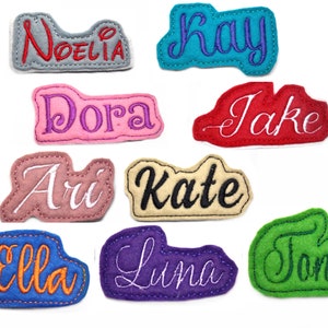 Puede incluir: Una colección de parches de nombres de fieltro de colores, en varios colores, incluyendo rojo, azul, rosa y verde. Cada parche presenta un nombre diferente bordado en un color contrastante, como "Noelia", "Kay", "Dora", "Jake", "Ari", "Kate", "Ella", "Luna" y "Toni".