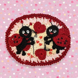 May include: An oval crochet wall hanging featuring two black ladybugs with red spotted wings on a cream background. The ladybugs hold strings attached to red crocheted hearts. The piece is framed by a dark red border with pink accents.