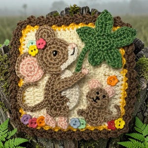 Può includere: Un arazzo quadrato all'uncinetto raffigurante un grande topo marrone che tiene in mano una foglia verde a cinque punte, accompagnato da un topo marrone più piccolo. Il pezzo è bordato in marrone scuro con finiture gialle e bottoni colorati.