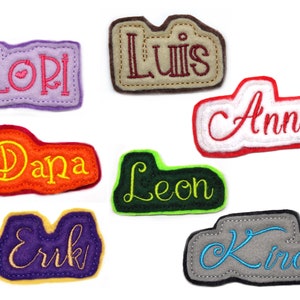 May include: A selection of felt name patches in various colours. Each patch displays a different name embroidered in colours such as pink, red, and blue, including Lori, Luis, Anna, Dana, Leon, Erik, and Kira. The patches have an irregular shape and are stitched.