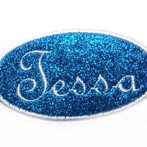 Könnte beinhalten: Ein blauer Glitzer-Aufnäher mit dem Namen "Tessa" in weißer Schrift gestickt. Der ovale Aufnäher mit weißem Rand eignet sich zum Personalisieren von Kleidung oder Accessoires.