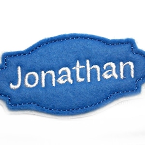 Könnte beinhalten: Ein blaues Filz-Namensschild mit dem Namen "Jonathan" in Weiß gestickt. Das Schild hat einen gewellten Rand und einen weißen Rand, wodurch ein kontrastreiches Design entsteht. Der Name ist auf dem Schild zentriert.