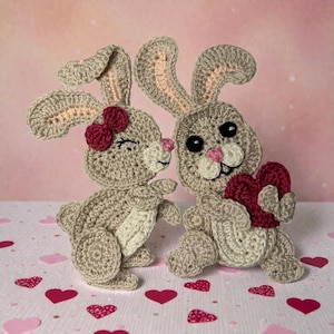 May include: Two crocheted bunny figures. One bunny has a red bow and is leaning in to kiss the other bunny. The second bunny is holding a red heart. The bunnies are light brown with pink and white accents. Red heart-shaped confetti is scattered around them.