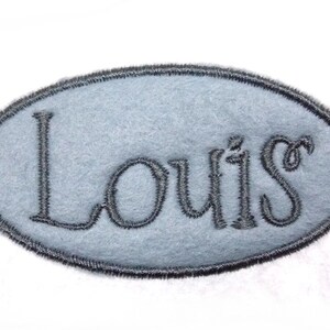 Könnte beinhalten: Ein hellblauer, ovaler Filz-Patch mit einem grauen gestickten Rand und dem Namen "Louis" in grauer Farbe.