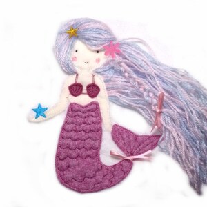 May include: A felt mermaid doll with long, purple and blue yarn hair. The mermaid is wearing a pink bikini top and a pink tail with a scale pattern. The doll is holding a blue star in its hand.