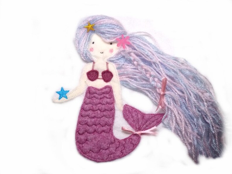 May include: A felt mermaid doll with long, purple and blue yarn hair. The mermaid is wearing a pink bikini top and a pink tail with a scale pattern. The doll is holding a blue star in its hand.