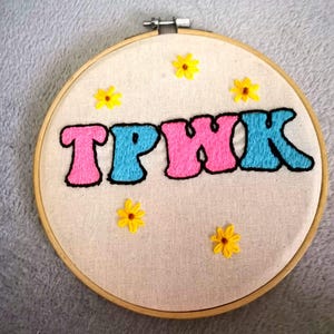 May include: Embroidered hoop art with the text "TPWK" in pink, blue, and black lettering. The background is white with yellow embroidered flowers.