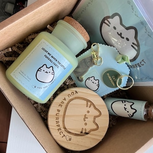 May include: A gift box containing cat-themed merchandise. It features a light green candle, a wooden coaster, a small vial, a keychain, and a notepad, all with a cartoon cat design. The box is filled with brown paper filler.
