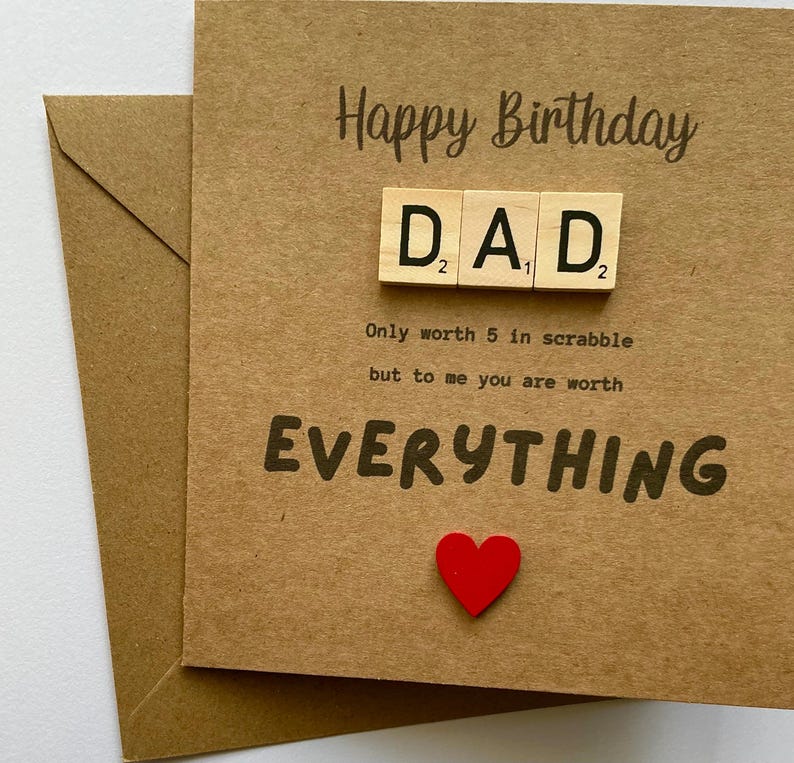 May include: A square brown kraft paper birthday card featuring the text Happy Birthday and DAD spelled out with wooden letter tiles. Below the tiles, the text reads Only worth 5 in scrabble but to me you are worth EVERYTHING, with a small red heart.