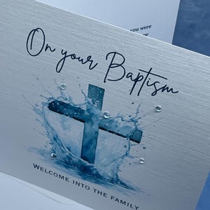 May include: A baptism card with a silver metallic finish. The card features a blue cross design with water splash effects and the words "On your Baptism" in elegant script. The bottom of the card reads "Welcome into the family."