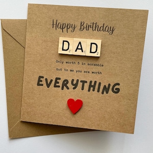 May include: A square brown kraft paper birthday card featuring the text Happy Birthday and three wooden Scrabble tiles spelling DAD. Below the tiles, the text reads Only worth 5 in scrabble but to me you are worth EVERYTHING, with a small red heart.