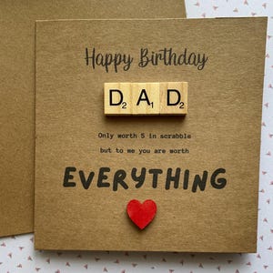May include: A brown kraft paper greeting card with the text Happy Birthday. Three wooden letter tiles spell DAD with numerical values. Below, the text reads Only worth 5 in scrabble but to me you are worth EVERYTHING, with a small red heart icon.
