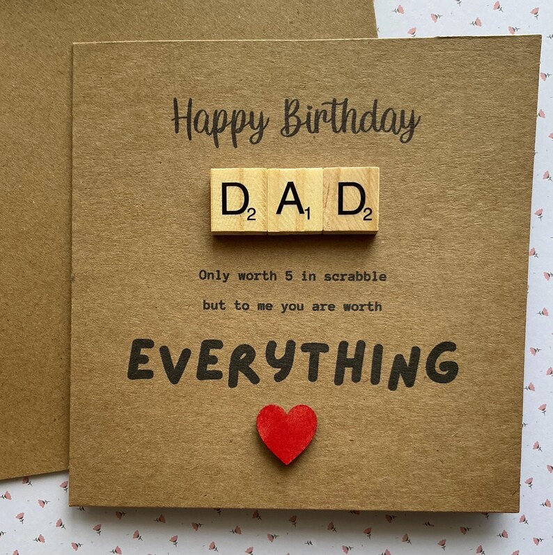 May include: A brown kraft paper greeting card with the text Happy Birthday. Three wooden letter tiles spell DAD with numerical values. Below, the text reads Only worth 5 in scrabble but to me you are worth EVERYTHING, with a small red heart icon.