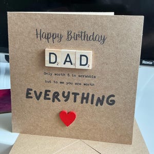 May include: A square brown kraft paper greeting card featuring the text Happy Birthday and DAD spelled out with three wooden letter tiles. Below the tiles, the text reads Only worth 5 in scrabble but to me you are worth EVERYTHING with a red heart.