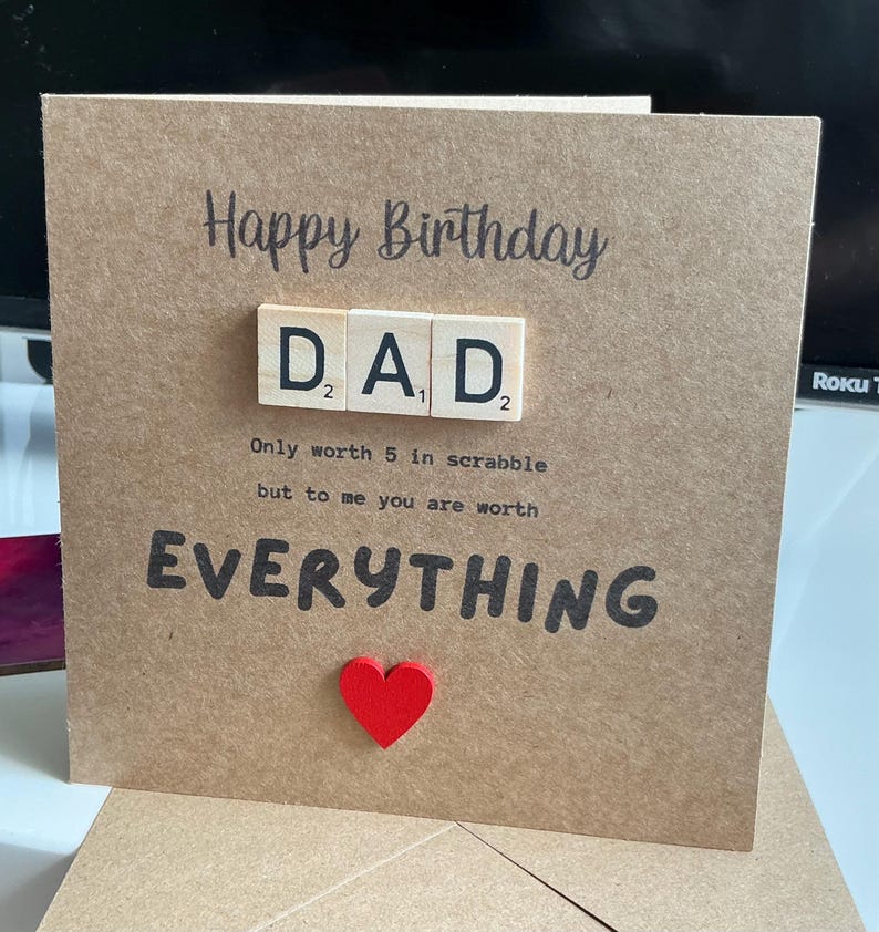 May include: A square brown kraft paper greeting card featuring the text Happy Birthday and DAD spelled out with three wooden letter tiles. Below the tiles, the text reads Only worth 5 in scrabble but to me you are worth EVERYTHING with a red heart.