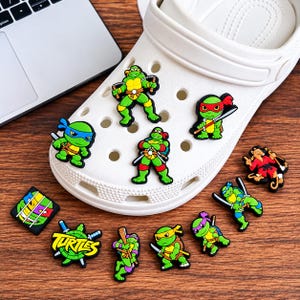 Peut inclure: Une chaussure sabot en mousse blanche décorée de plusieurs breloques colorées représentant les personnages des Tortues Ninja, incluant diverses tortues en action, un rat, et un carré avec le mot Turtles écrit en jaune.