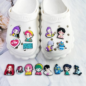 May include: White clogs adorned with various colorful K-Pop-themed charms. The charms feature cartoon characters and the phrases "K POP" and "K-POP DEMON HUNTERS". The charms are in various shapes and sizes.