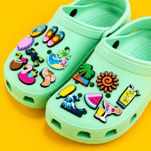 May include: A pair of light mint green foam clogs decorated with various colourful summer-themed charms, including a sun, palm tree, watermelon slice, ocean wave, flip-flops, a cocktail, a flamingo float, and a scuba diver on the perforated surface.