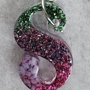 May include: A clear resin letter "S" keychain with glitter accents. The top of the "S" is green, transitioning to pink and purple at the bottom. A silver-colored clasp is attached for easy use.