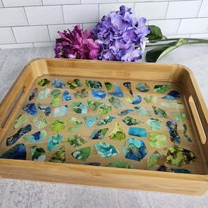 May include: A rectangular bamboo serving tray with a clear resin surface. The resin is embedded with colorful abstract shapes in shades of blue, green, and white. The tray has built-in handles.