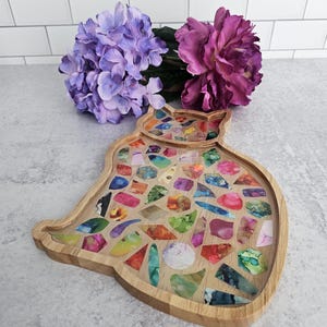May include: A wooden cat-shaped tray filled with colorful, irregularly shaped pieces. The tray has a natural wood finish and is filled with a mosaic of vibrant colors. Purple and pink flowers are in the background.