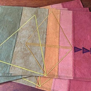 May include: Set of four placemats with a geometric design. The placemats are made of a natural fabric and have a patchwork of different colors, including green, brown, orange, and pink. The design features a yellow diamond shape with a smaller diamond shape inside. The placemats have a purple triangle design on the bottom right corner.