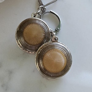May include: A pair of silver tone earrings with a round, cream-colored button-like design. The earrings have a simple, elegant style.