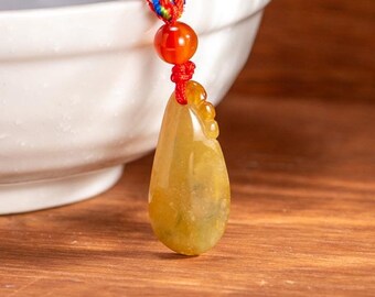 Puede incluir: Un colgante de jade amarillo con una cuerda roja y cuentas. El colgante tiene forma de lágrima y tiene una superficie lisa y pulida. El colgante está colgado de un cuenco blanco.