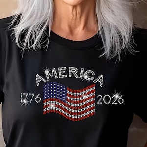 Woman wearing black rhinestone America 250th t-shirt with red, white, and blue flag, smiling against brick wall.