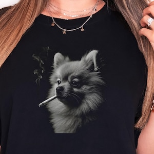 May include: Black t-shirt featuring a grayscale illustration of a Pomeranian dog smoking a cigarette. The dog is depicted with detailed fur and a contemplative expression, with smoke rising from the cigarette. The shirt is a casual wear item.