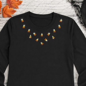 May include: A black long-sleeve shirt featuring a decorative neckline pattern made of small, triangular candy corn-shaped rhinestones in yellow, orange, and white. The shirt is paired with dark blue denim jeans, creating a simple, casual look.