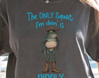 May include: Dark gray t-shirt featuring a cartoon frog wearing a cowboy hat, boots, and a gun. The text above the frog reads "The ONLY Squat I'm doin' is" and below the frog, the text reads "DIDDLY".