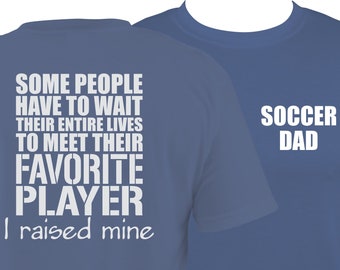 Puede incluir: Dos camisetas azules con texto blanco. La camiseta delantera dice "Soccer Dad". La camiseta trasera dice "Algunas personas tienen que esperar toda su vida para conocer a su jugador favorito. Yo crié al mío."