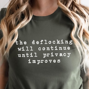 May include: An olive green short-sleeved t-shirt featuring white typewriter-style text on the front that reads: the deflocking will continue until privacy improves. The shirt has a crew neckline and is tied in a knot at the bottom center.
