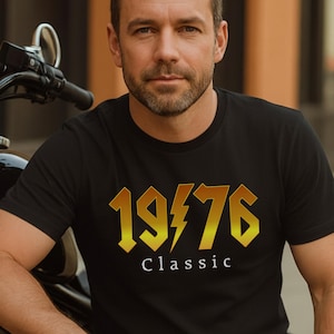 May include: Black t-shirt with the text "1976 Classic" in gold lettering. The design includes a lightning bolt graphic. The shirt is worn by a person sitting next to a black motorcycle.