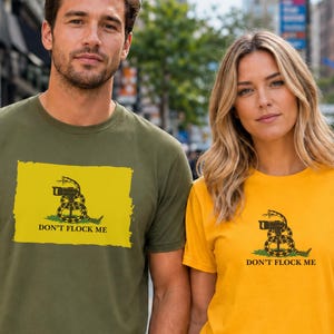 May include: An olive green t-shirt and a yellow t-shirt, each featuring a graphic of a coiled goose with the text DON'T FLOCK ME. The graphic depicts a goose in a defensive posture on a patch of green grass, mimicking a historical flag design.