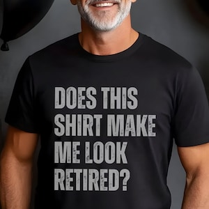 May include: A black short-sleeved t-shirt featuring the text "DOES THIS SHIRT MAKE ME LOOK RETIRED?" printed in a distressed, light gray, sans-serif font across the chest. The garment has a classic crew neckline and a standard fit.
