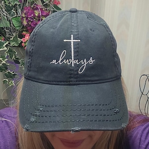 May include: A distressed navy blue baseball cap with the word "always" embroidered in white script below a white cross. The cap has a pre-curved bill and distressed detailing.