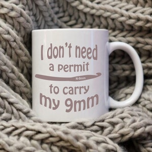 May include: White ceramic mug with a brown graphic that says "I don't need a permit to carry my 9mm" with a small crochet hook graphic and the text "N-9mm" above the hook.