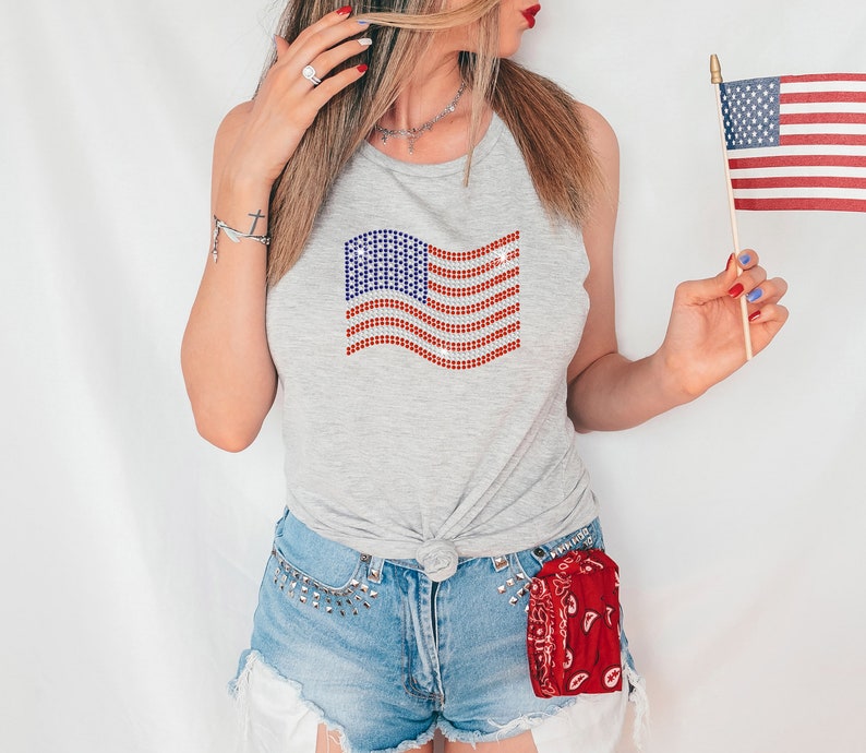 May include: A woman wearing a gray tank top with a red, white, and blue sequined American flag design. She is also wearing denim shorts with a red bandana tucked into the waistband. She is holding a small American flag on a stick.