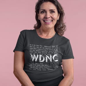 Może przedstawiać: Ciemnoszara koszulka z napisem "WDNC" dużymi białymi literami. Koszulka zawiera r&oacute;wnież mniejszy biały tekst z frazami takimi jak "We Do Not Care Club" i "Menopausal Women Unite".