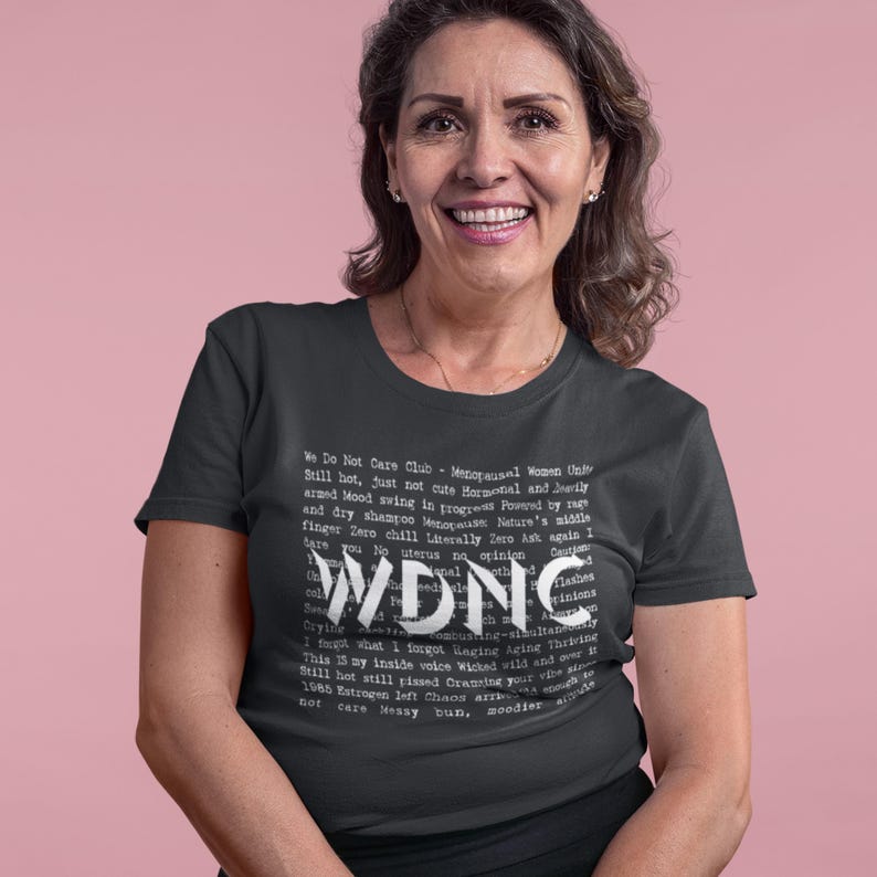 Może przedstawiać: Ciemnoszara koszulka z napisem "WDNC" dużymi białymi literami. Koszulka zawiera r&oacute;wnież mniejszy biały tekst z frazami takimi jak "We Do Not Care Club" i "Menopausal Women Unite".